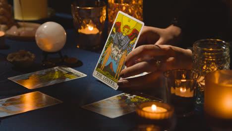 Close-Up-Of-Woman-Giving-Tarot-Card-Reading-On-Candlelit-Table-Holding-The-Lovers-Card